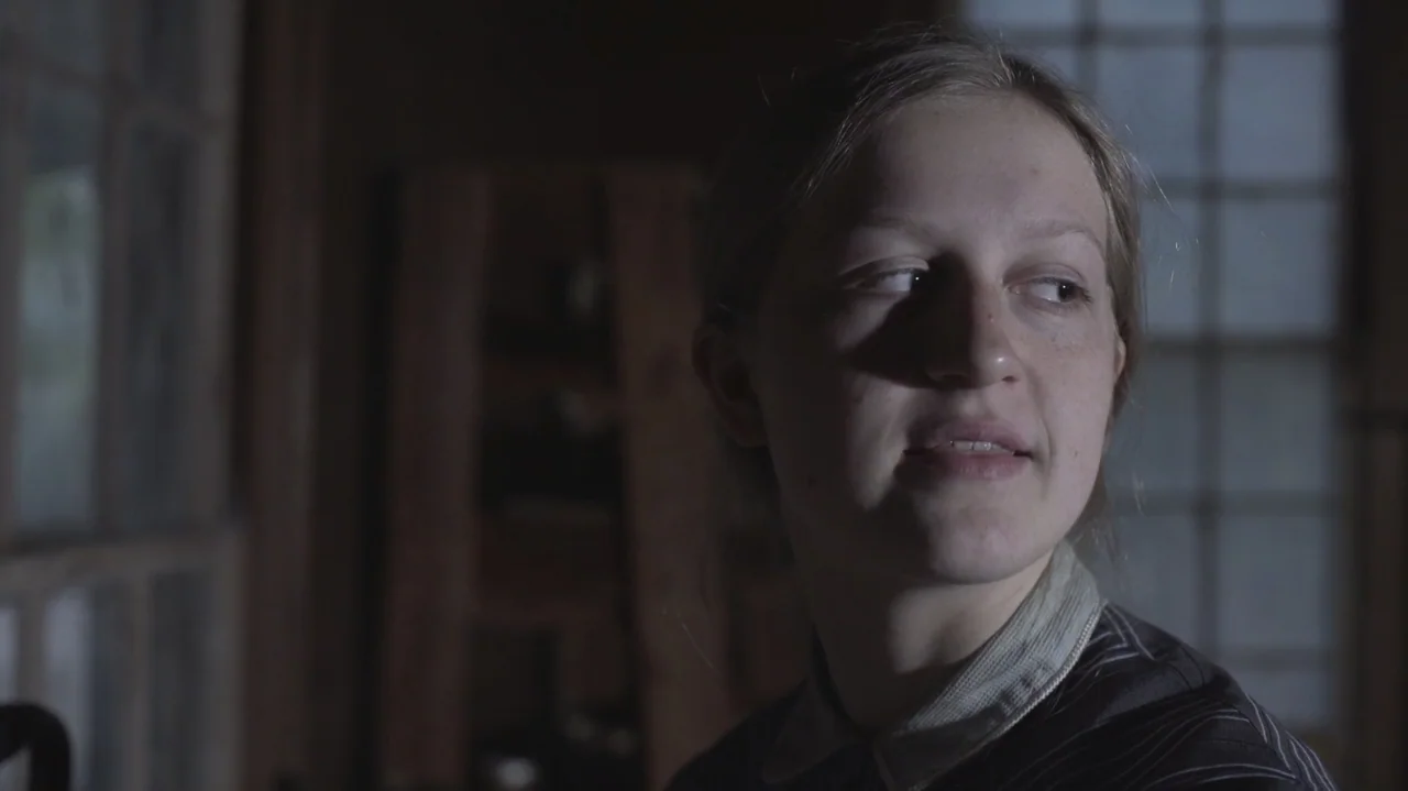 The Dalton Gang (2020) movie still: closeup — A dimly lit room with a paneled window in the background and wood walls. A woman in a dark shirt turns…