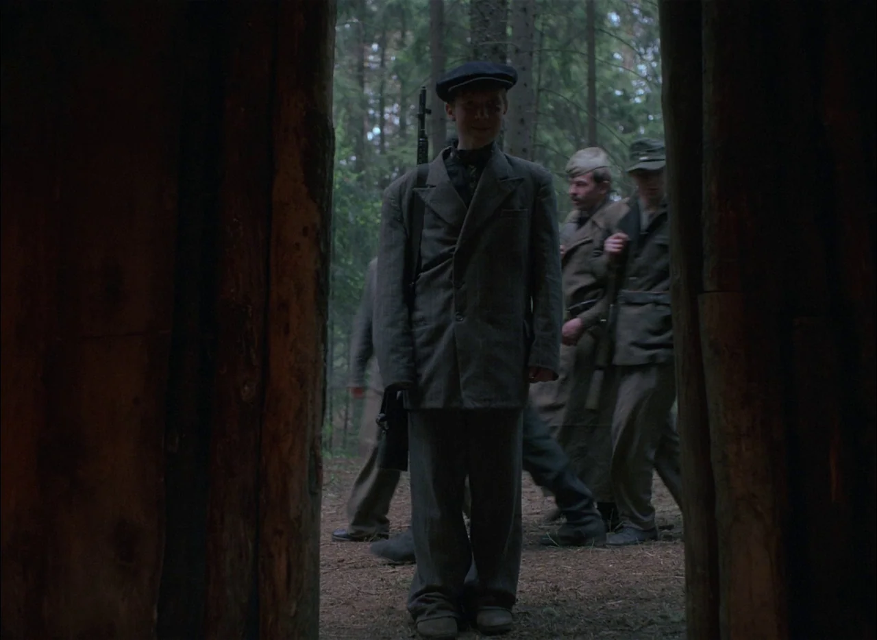 Come and See (1985) movie still: fullbody — A teenage boy in a wool suit stands in a dark wooded area. Soldiers walk in the background through the…