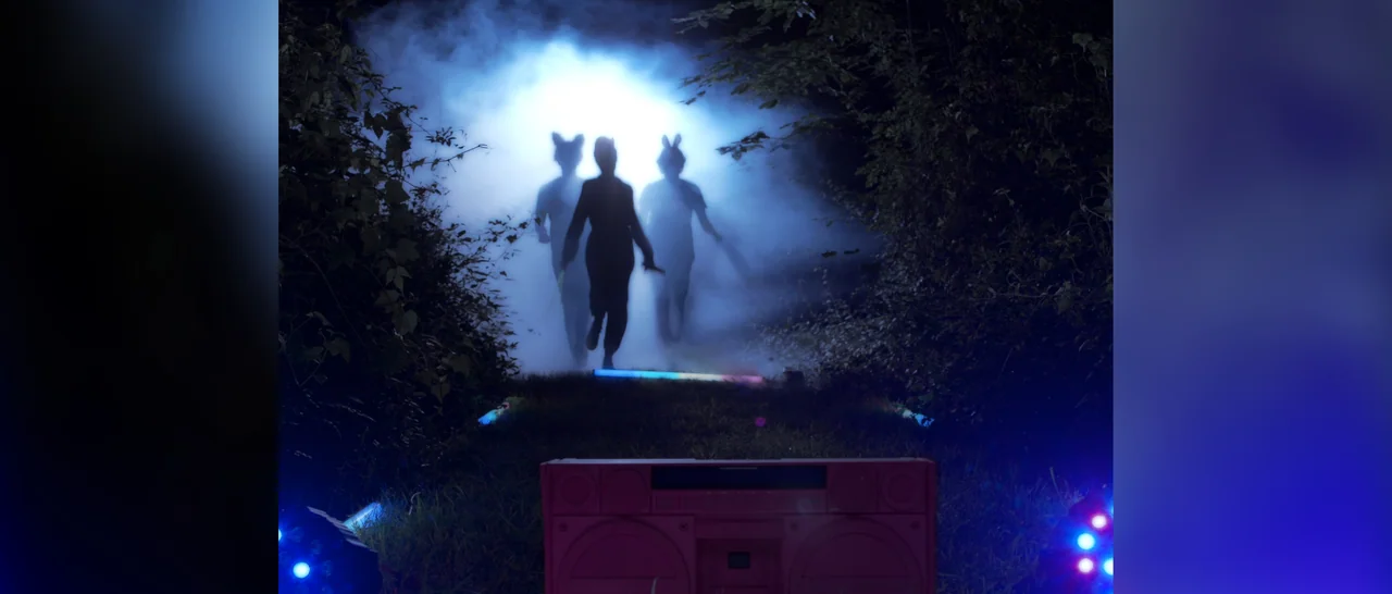 Songs For A Sloth (2021) movie still: wide — Three silhouetted figures emerge from thick blue-tinted fog in a dark grassy field. A vintage pink…