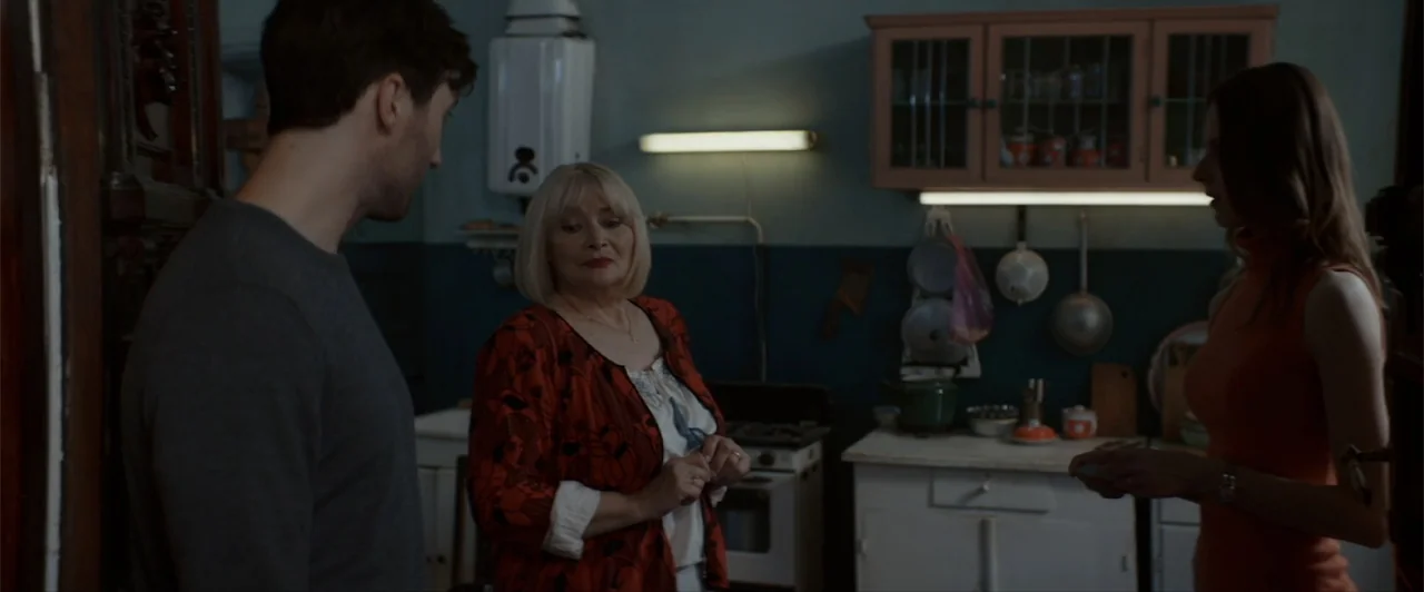 La herencia (2010) movie still: medium — A modest home kitchen features vintage cabinets, a gas stove, and utilitarian wall decor. Three individuals…