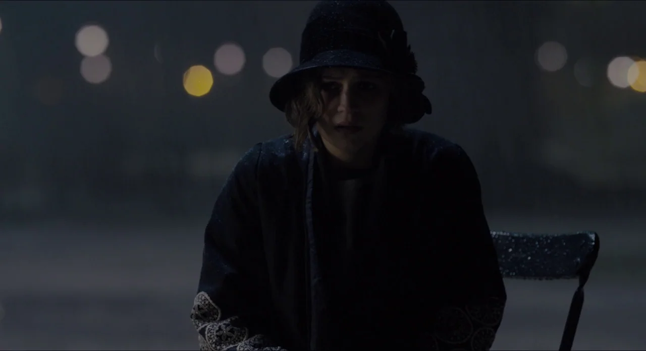 The Danish Girl (2015) movie still: medium — A woman sits on a metal chair at night in an outdoor space. Distant out-of-focus street lights create a…