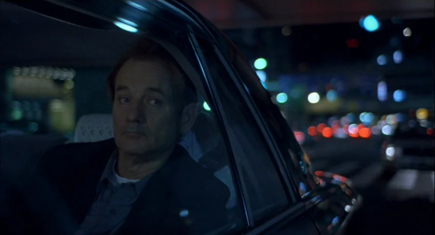 Lost In Translation (2003) movie still: medium — A man sits in the back of a taxi at night. City traffic lights blur in the dark background outside…