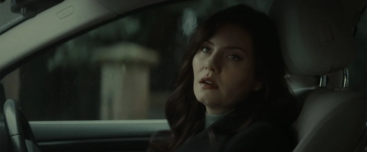 The Cellar (2022) movie still: medium — An interior of a modern car during a rainy or overcast day. A woman sits in the passenger seat looking…