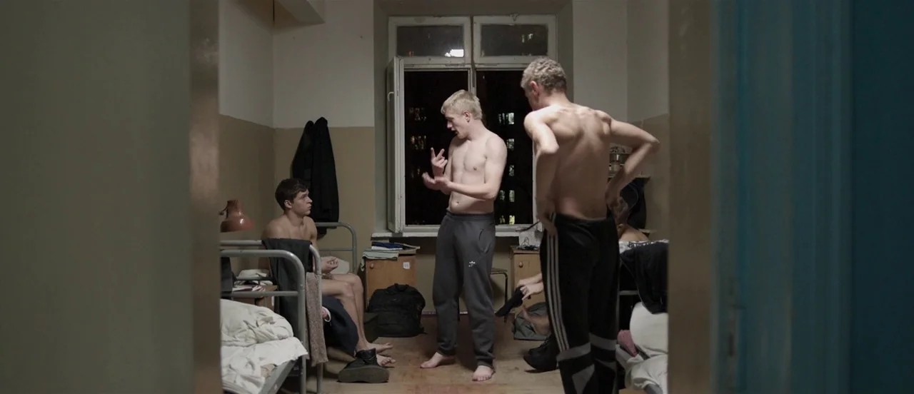 The Tribe (2014) movie still: medium — A cluttered shared living space with metal bunk beds and a central window. Three young men interact in the…