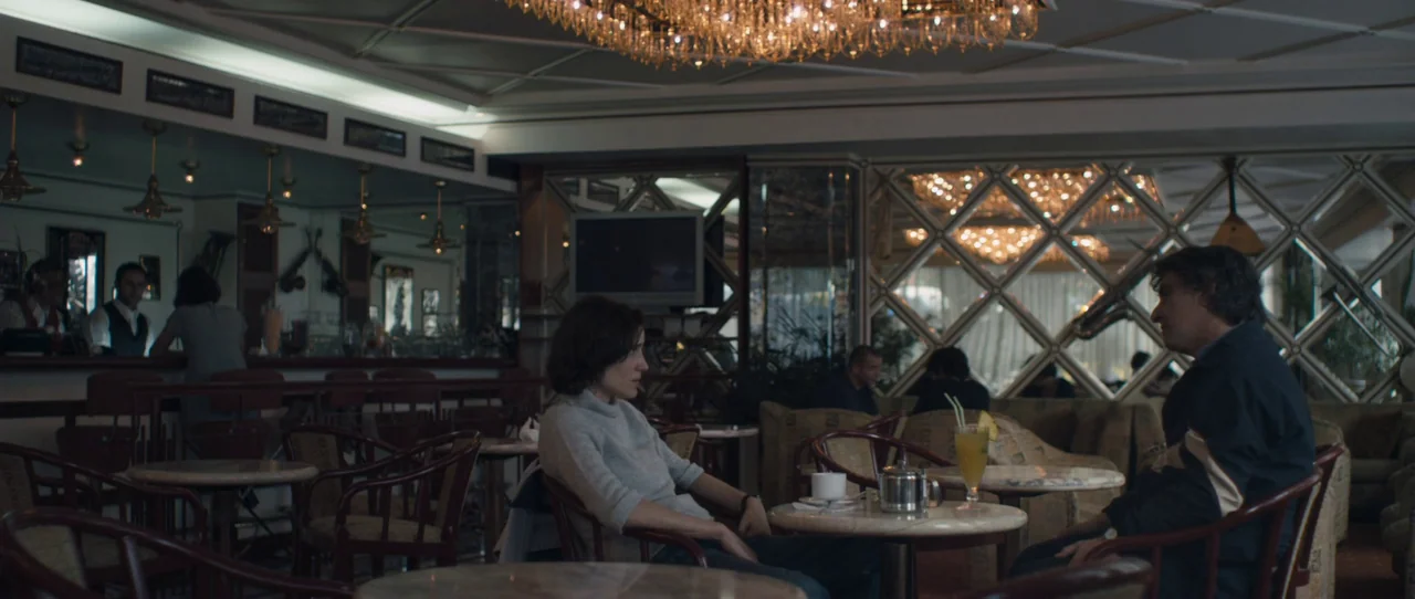 Alps (2011) movie still: medium — A dimly lit hotel bar with diamond-patterned mirrored walls and ornate crystal chandeliers. A woman and a man are…