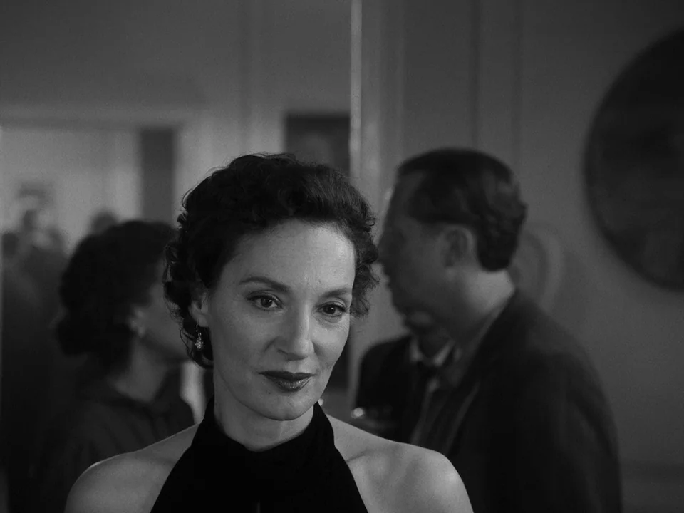Cold War (2018) movie still: medium — A woman stands in a social setting with people mingling in the blurred background. She faces forward while…
