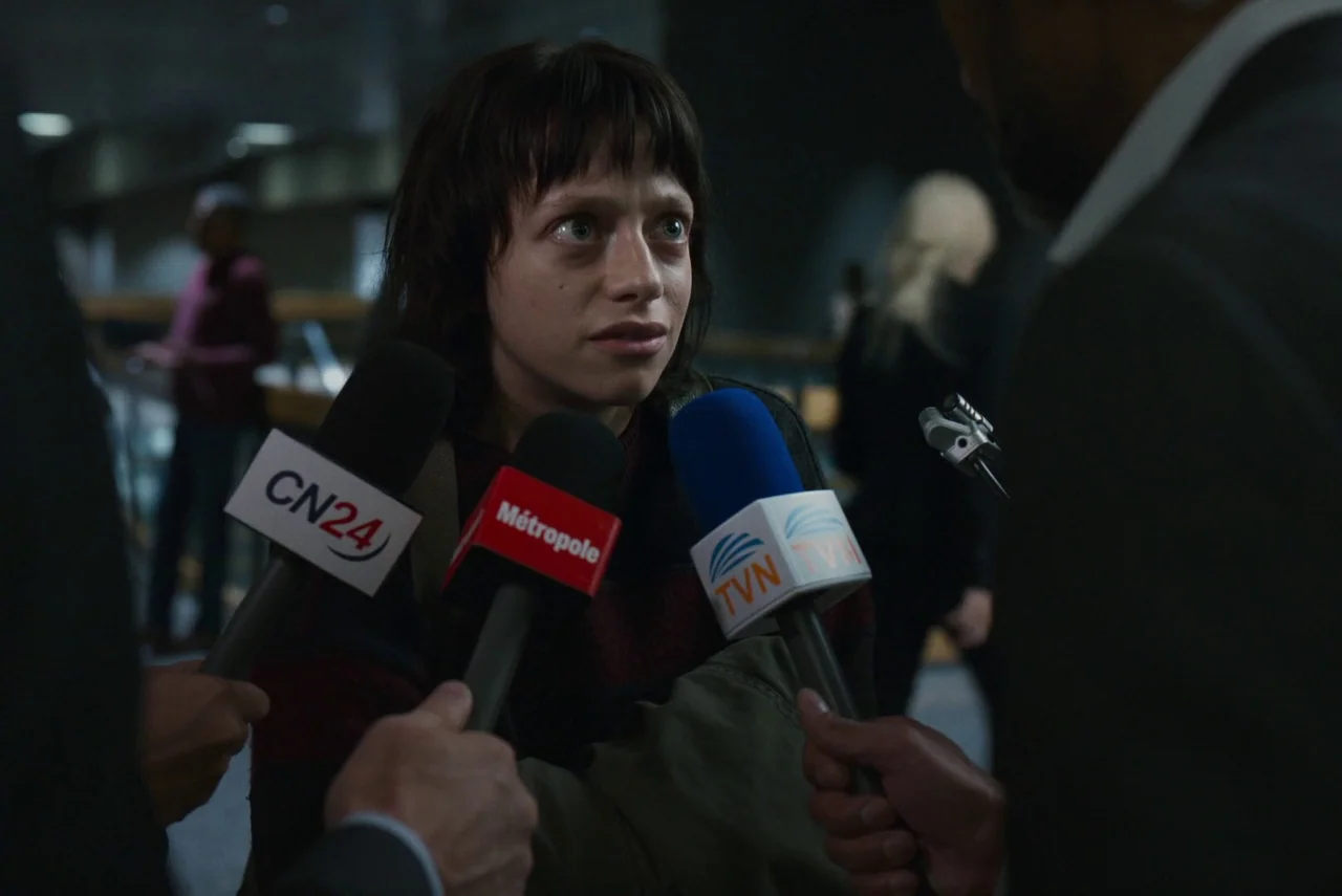 Red Rooms (2023) movie still: medium — A dimly lit indoor space crowded with people holding microphones toward a central figure. A young woman…