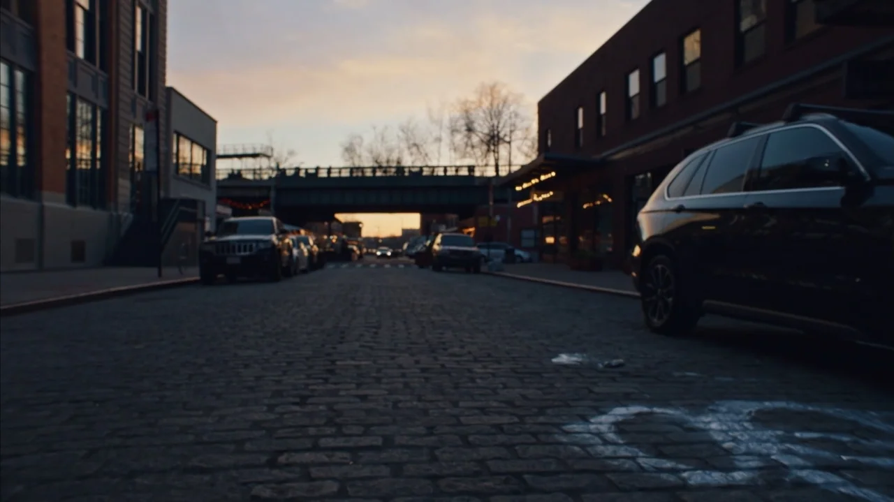 The Stroll (2023) movie still: establishing — A quiet cobblestone street is lined with multi-story brick buildings under a twilight sky. Several…