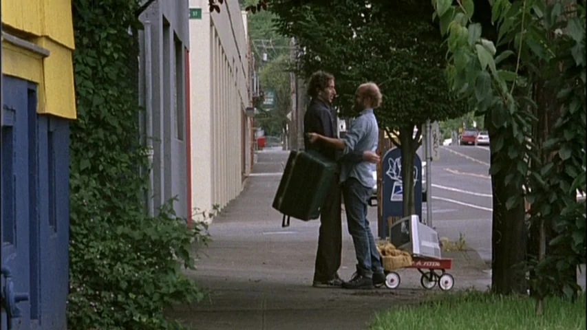 Old Joy (2006) movie still: fullbody — A quiet urban sidewalk lines a building covered in creeping vines and blue paint. Two men stand on the…