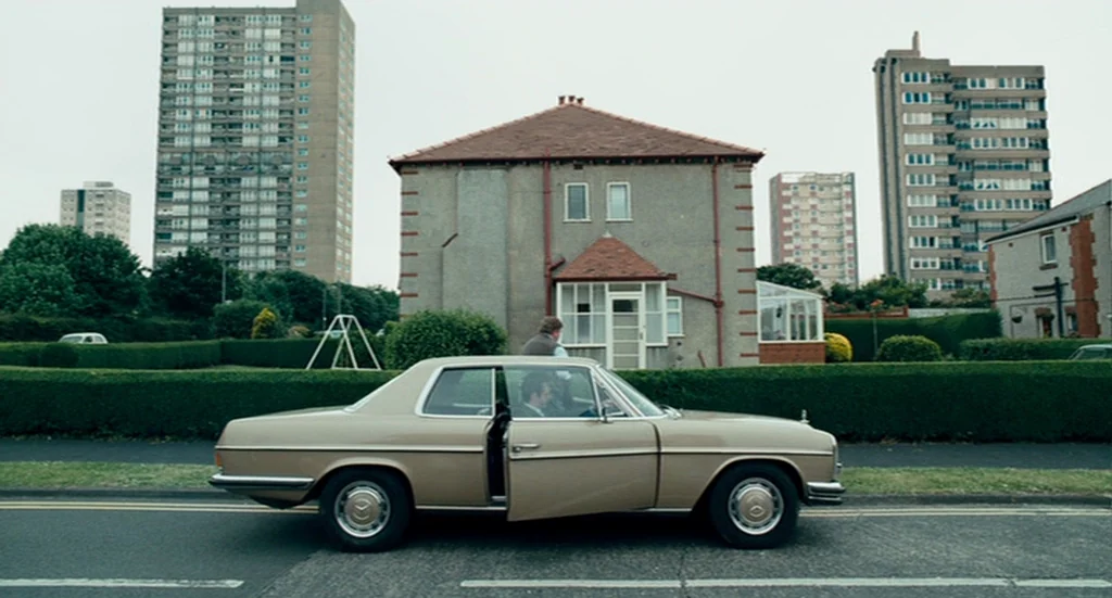 The Damned United (2009) movie still: wide — A suburban residential street features a beige car parked in front of a house. Tall concrete apartment…