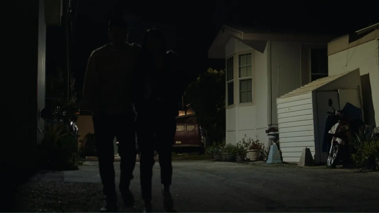 The Cleaner (2021) movie still: fullbody — A night view in a residential mobile home park with white structures and vehicles. Two people walk…