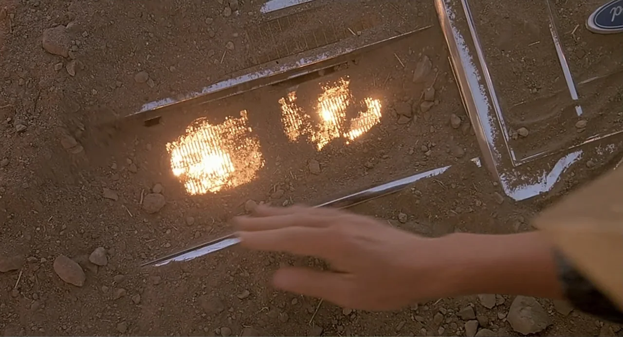 Tremors (1990) movie still: closeup — A buried vehicle front end sits embedded in dry cracked desert soil. A human hand extends into the frame…