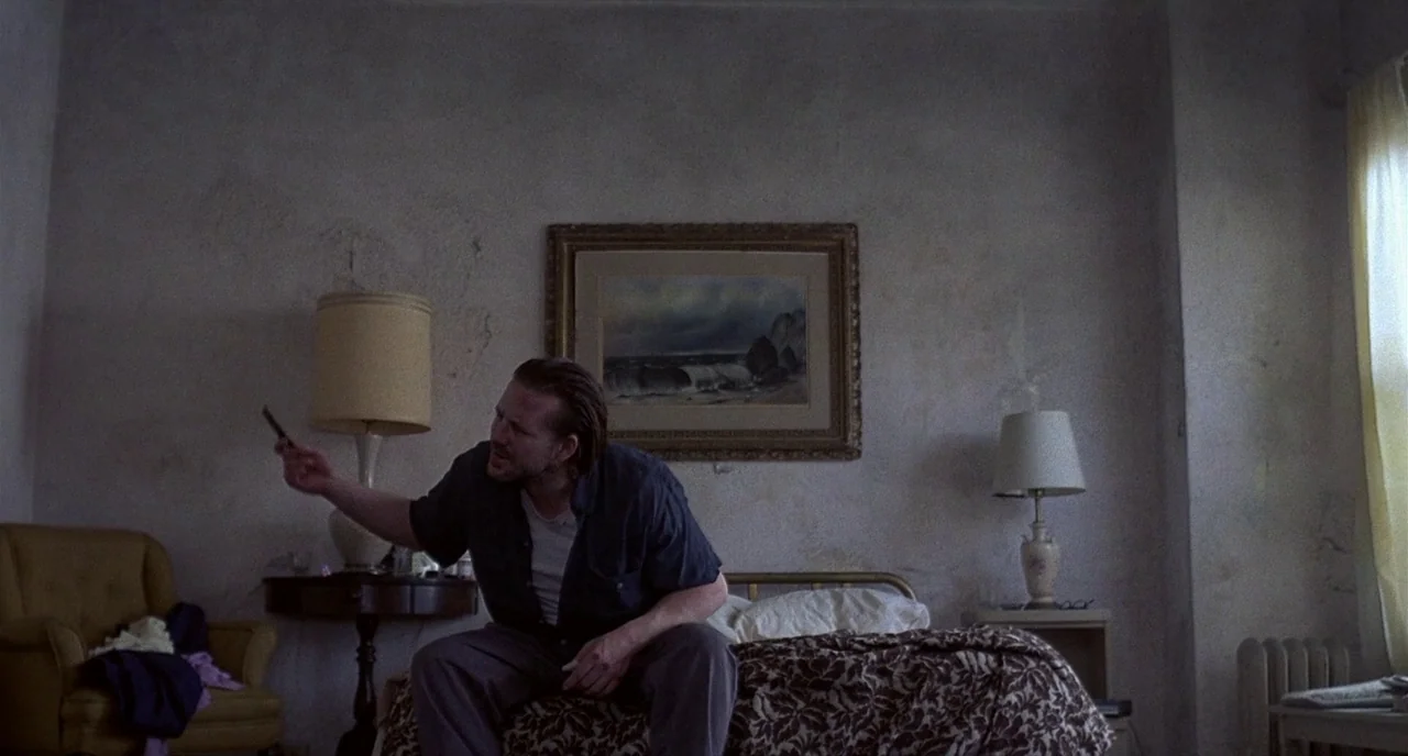 Barfly (1987) movie still: medium — A dated motel room features peeling walls decorated with a single framed landscape painting. A man sits on the…