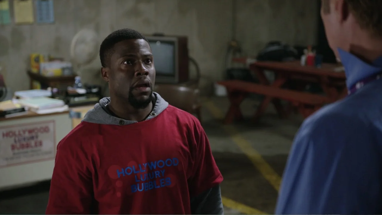 Get Hard (2015) movie still: medium — A cluttered concrete garage features a picnic table and an old television in back. A man stands in the…