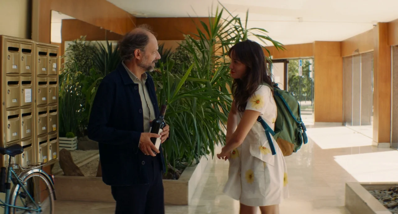 Anaïs in Love (2021) movie still: medium — A sunlit apartment building lobby features modern wood paneling and large decorative indoor palms. A…