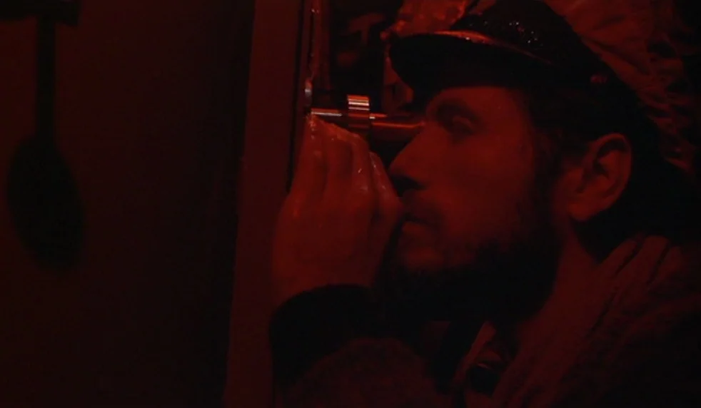Das Boot (1981) movie still: closeup — A man peers intently through a small metal peephole in a darkened space. The environment is submerged in deep…