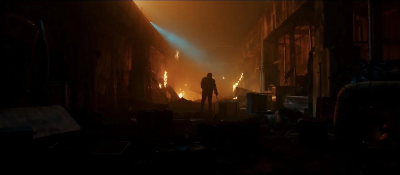 Guns Akimbo (2020) movie still: wide — A dark, expansive industrial hall is filled with smoke and active, flickering fire. A lone man stands in the…