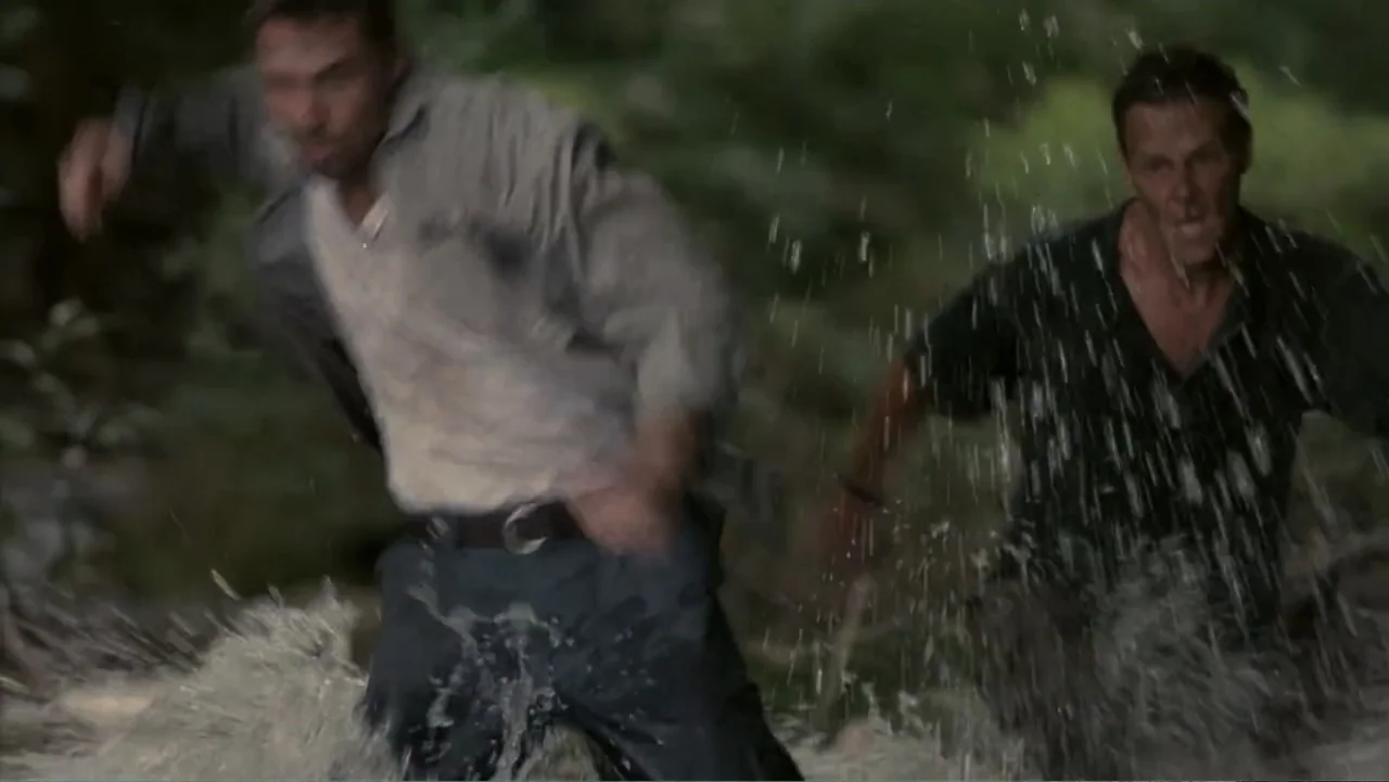 Two men are running through a shallow, dark forest river. Water sprays around their legs as they move toward the camera.