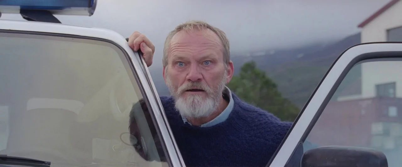 A White, White Day (2019) movie still: medium — An older man leans from the driver's side of a vehicle in an overcast environment. Green hills and a…
