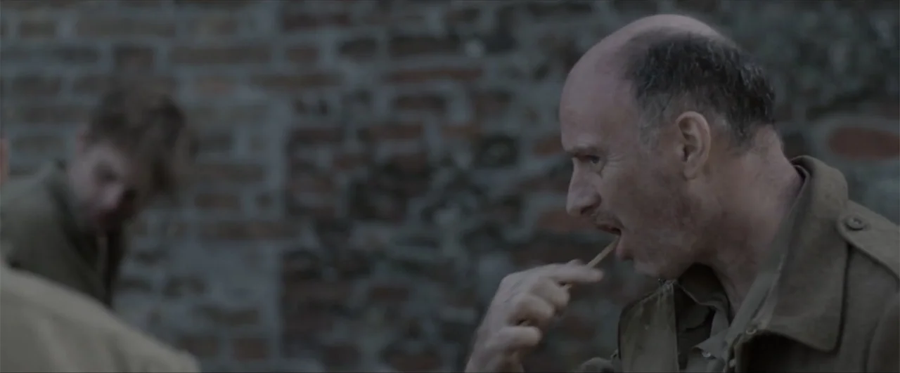 Behind the Line: Escape to Dunkirk (2020) movie still: medium — An outdoor area features a blurred brick wall background with two men standing…