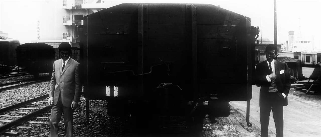 Tokyo Drifter (1966) movie still: wide — An industrial railway yard features gravel tracks and a large, dark wooden freight car. Two men in formal…