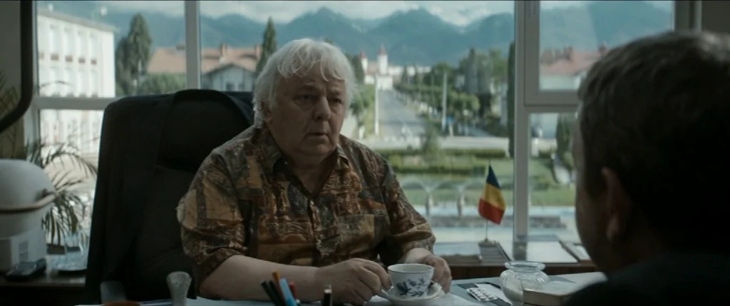 Graduation (2016) movie still: medium — An office features a large window overlooking a street and distant mountains. An elderly man sits at his…