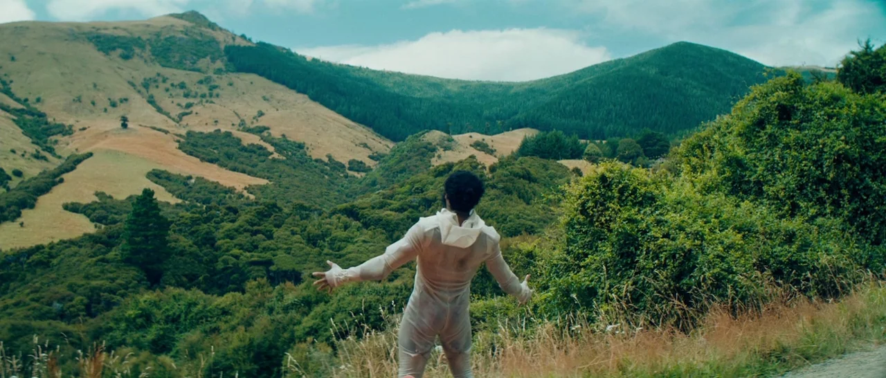 Z for Zachariah (2015) movie still: fullbody — A vast hilly terrain features dense green forests and patches of dry grass. A man in a white jumpsuit…