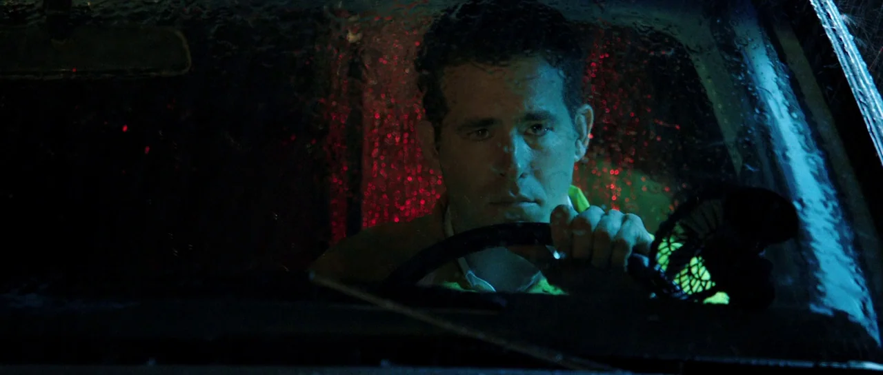 The Voices (2014) movie still: closeup — A dark car interior at night with rain droplets visible on the windshield. A man looks ahead while gripping…