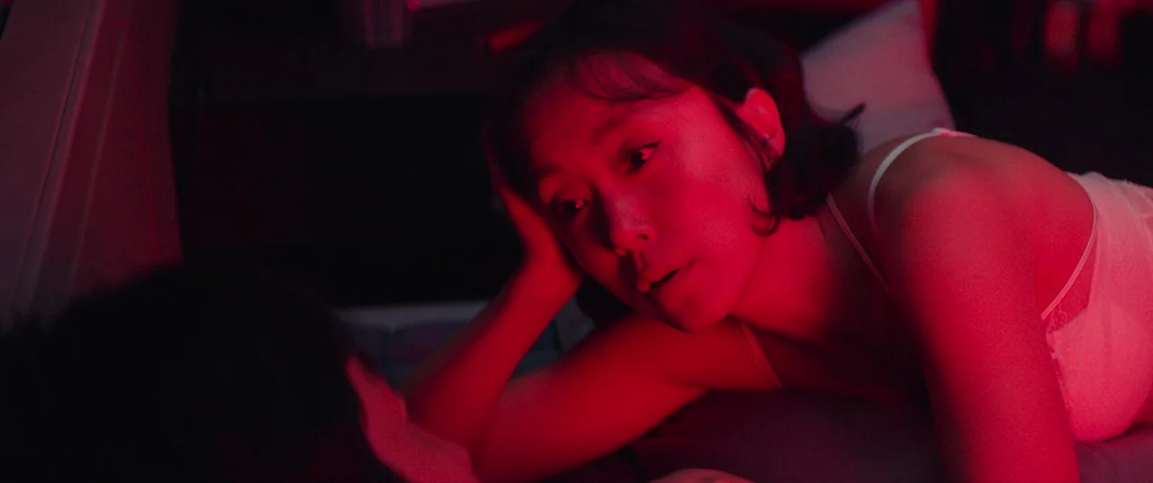 Beasts Clawing At Straws (2020) movie still: closeup — A dark bedroom is lit by a prominent red light source. A woman lies propped on her elbow…