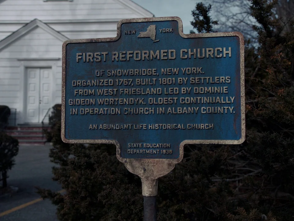 First Reformed (2017) movie still: closeup — A blue metal historical plaque stands on a post in front of a building. The text on the plaque details…