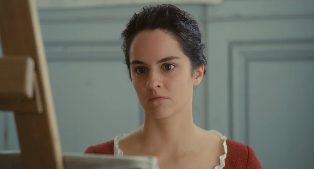 Portrait of a Lady on Fire movie still: closeup — A woman stands in a bright room with grey paneled walls. She gazes intently toward an easel…