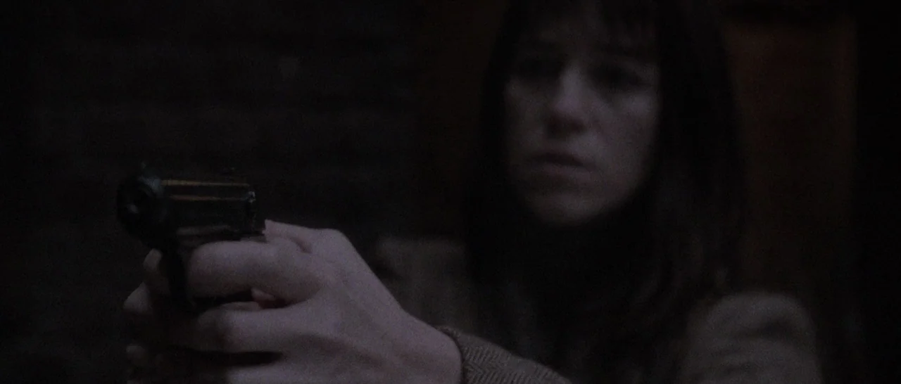 Nymphomaniac Vol 2 (2013) movie still: medium — A dark, dimly lit indoor space with indistinct background textures. A woman holds a handgun leveled…