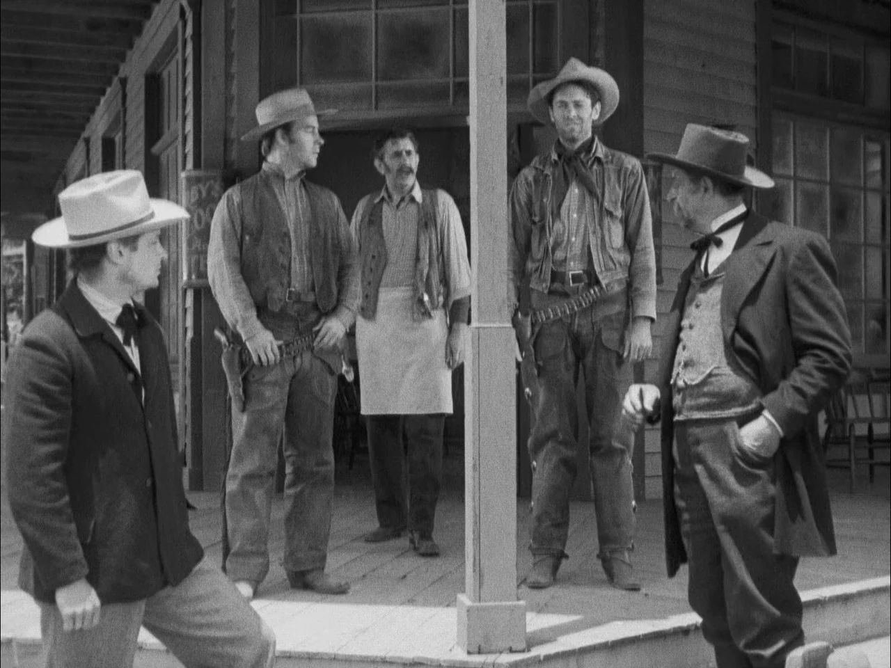 The Ox-Bow Incident (1942) movie still: fullbody — A wooden porch outside a building features five men gathered in casual conversation. The men wear…