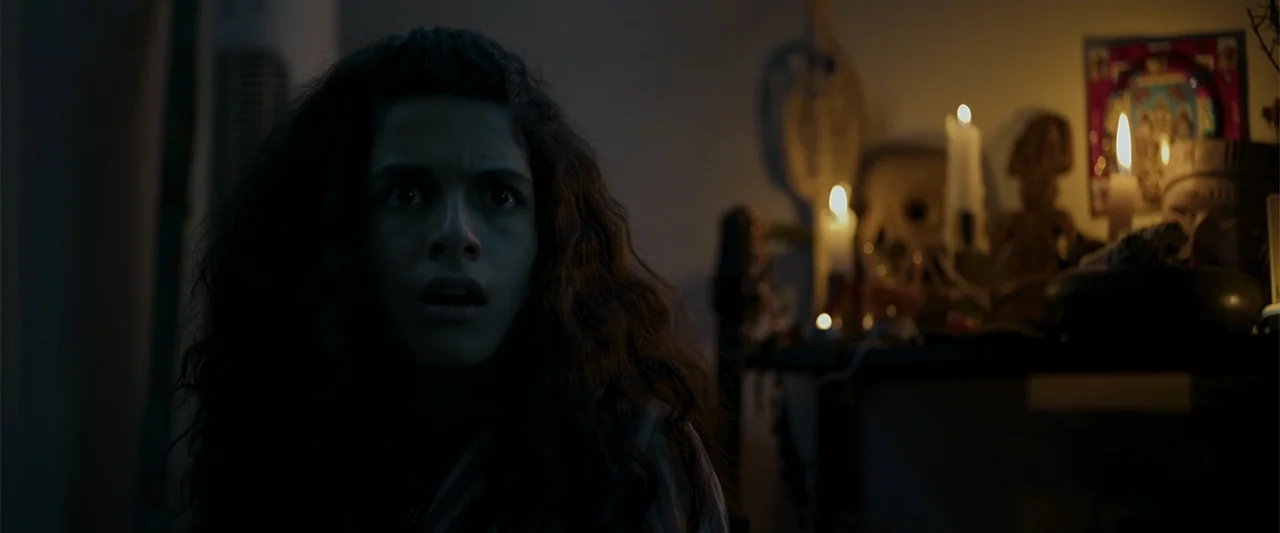 Evil Eye (2020) movie still: closeup — A dimly lit room features a small table with flickering candles and ritual objects. A young woman with long…
