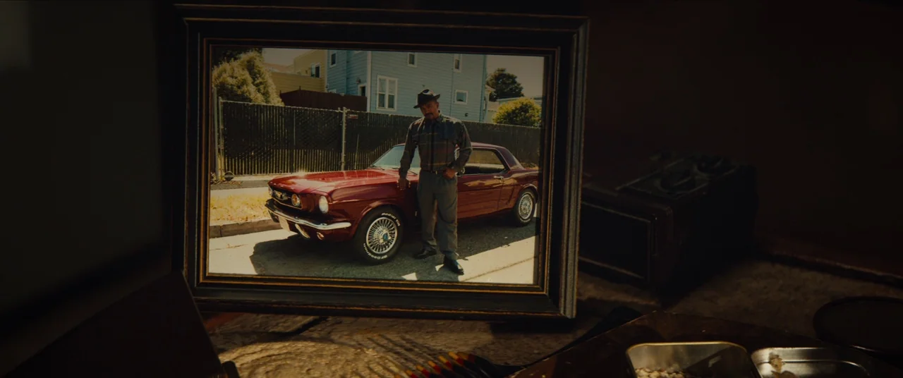 Sorry to Bother You (2018) movie still: closeup — A framed photograph sits on a dark surface inside a dimly lit room. The photo shows a man standing…