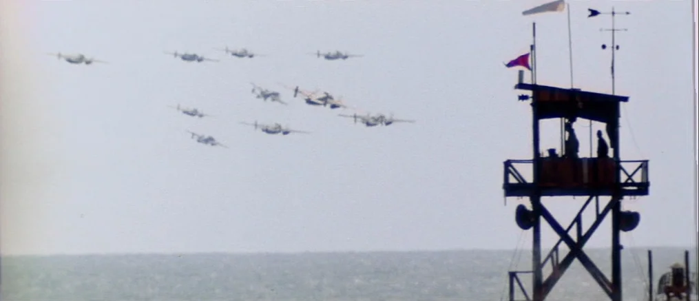 Catch 22 (1970) movie still: wide — A wooden observation tower stands at the edge of a flat, gray ocean. Multiple propeller-driven airplanes fly in…
