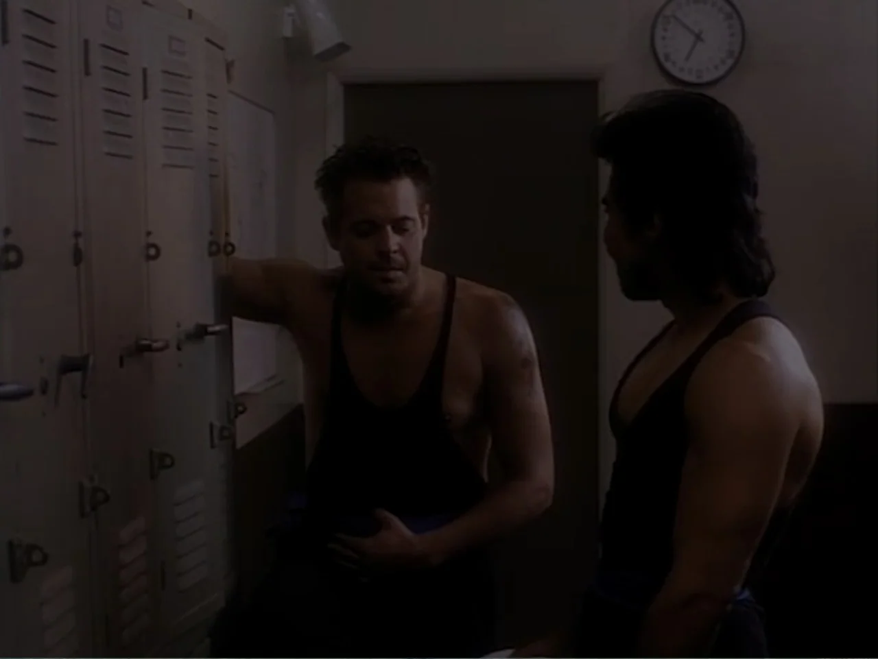 Firepower (1979) movie still: medium — A dimly lit locker room features rows of metal lockers and a wall clock. Two athletic men wearing matching…