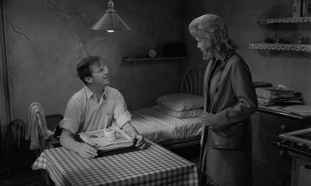 Something Wild (1961) movie still: medium — A spartan room features a small table with a checked cloth and a bed. A man with a bandaged eye sits…