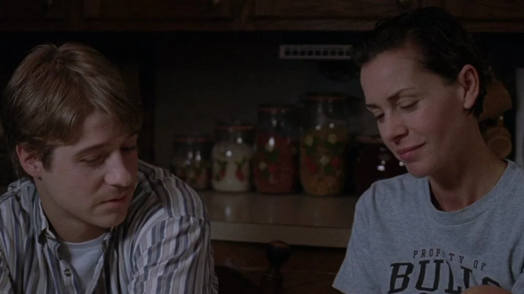 Junebug (2005) movie still: medium — A dark kitchen features glass jars of ingredients on the counter in the background. A young man and woman sit…