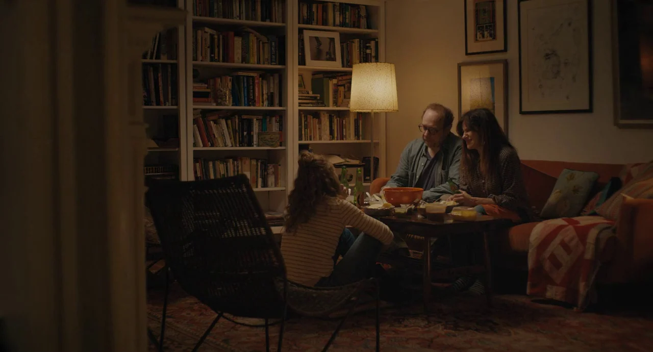 Private Life (2018) movie still: wide — A cluttered domestic room features floor-to-ceiling bookshelves lining the walls behind a sofa. Three people…