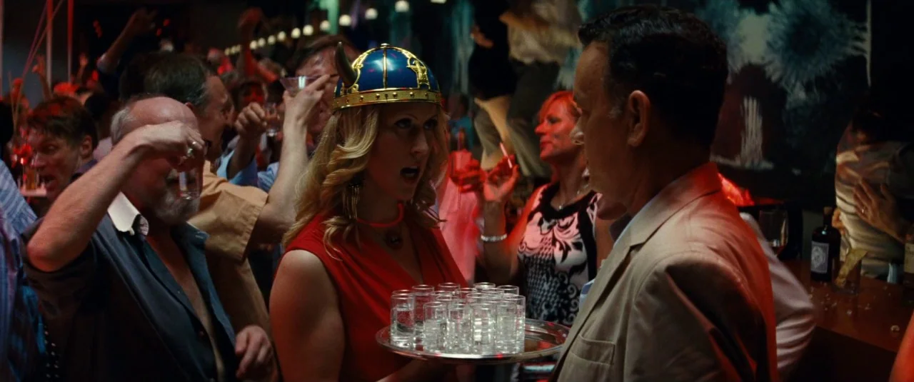 A Hologram for the King movie still: medium — A crowded bar setting with people holding drinks in a low-light indoor environment. A woman in a…