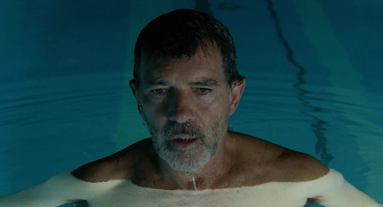 Pain and Glory (2019) movie still: closeup — A man stands chest-deep in a dark blue indoor swimming pool at night. He stares ahead while the water…