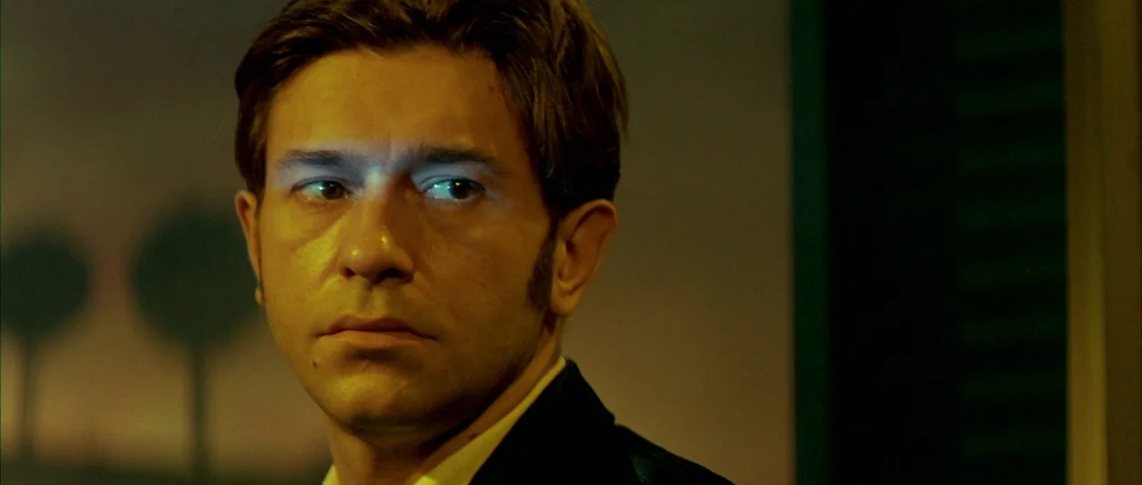 Querelle (1982) movie still: closeup — An interior space is dimly lit with artificial blue and warm yellow light sources. A man in a dark suit looks…