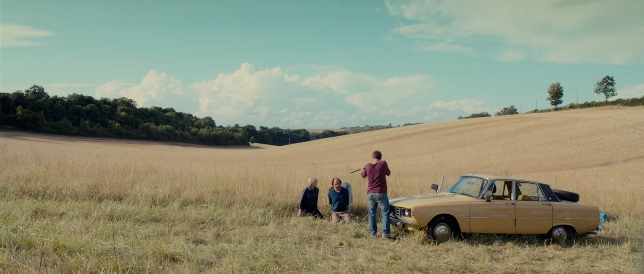 Road Games (2015) movie still: wide — A vast, dry grassy field stretches toward a treeline under a bright blue sky. One man stands pointing a rifle…