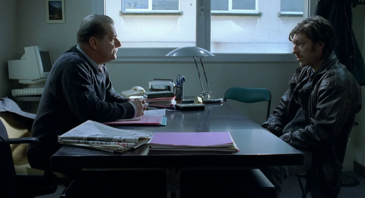 Read My Lips (2001) movie still: medium — A spartan office with a metal desk, stacks of folders, and a vintage computer monitor. Two men face each…
