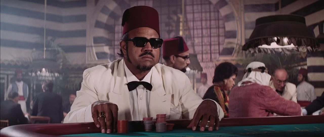Diamonds Are Forever (1971) movie still: medium — A dimly lit casino interior features patrons sitting at a green-felt gambling table. A man wearing…