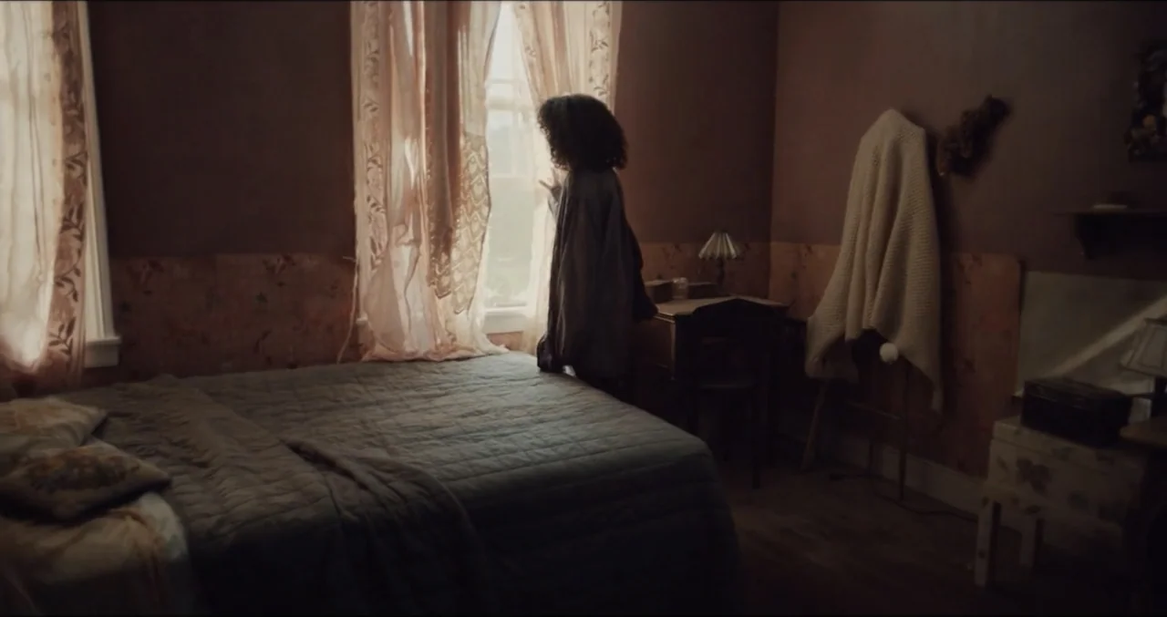 Eye for an Eye (2025) movie still: medium — A dimly lit bedroom features patterned wallpaper and a quilt-covered bed near a window. A person with…