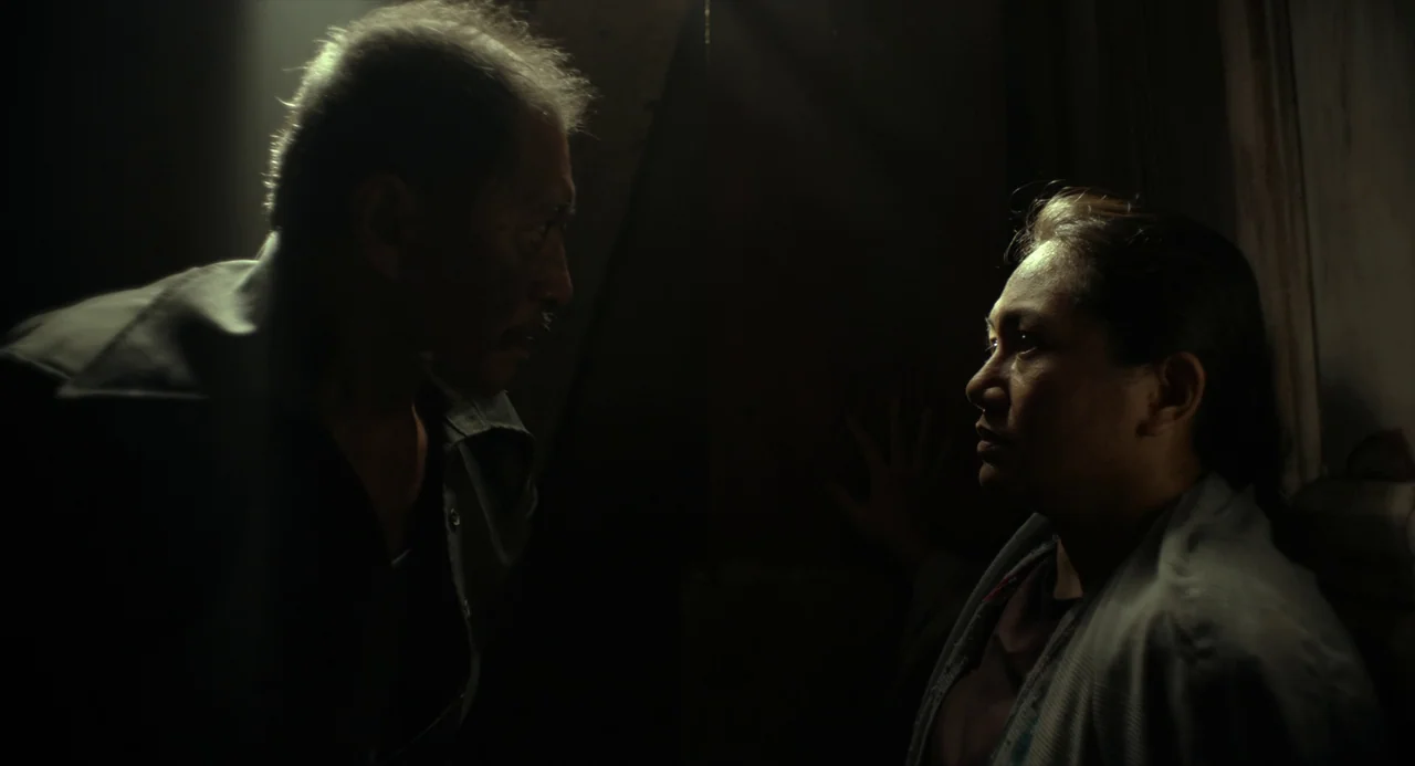 In My Mother’s Skin (2023) movie still: medium — A dark, minimally lit room serves as the setting for the two individuals. The man stands on the…