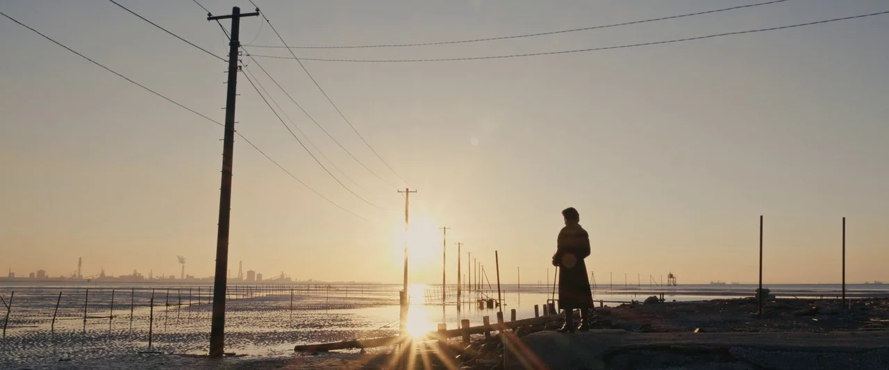 Shin Kamen Rider (2023) movie still: wide — A flat coastal tideland stretches toward a distant industrial horizon at sunset. A lone woman in a long…