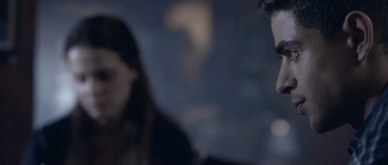 The Lullaby (2019) movie still: closeup — An interior space with a man in the foreground and a blurred woman behind. The man stares across the frame…